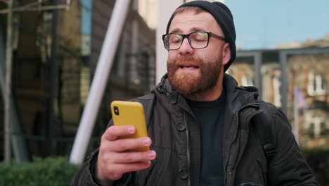 bearded man reacting and talking on smartphone with coffee in city