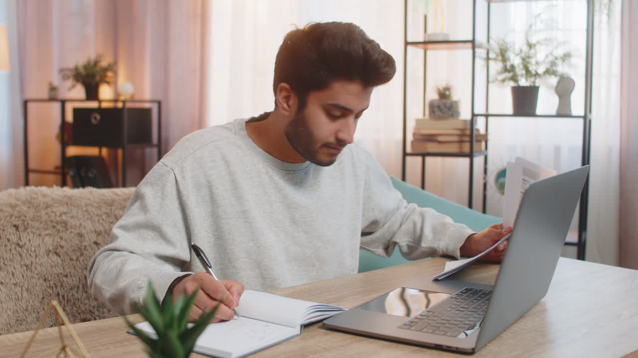 Giovane libero professionista indiano che esamina i report finanziari sul laptop mentre prende appunti nel suo ufficio domestico