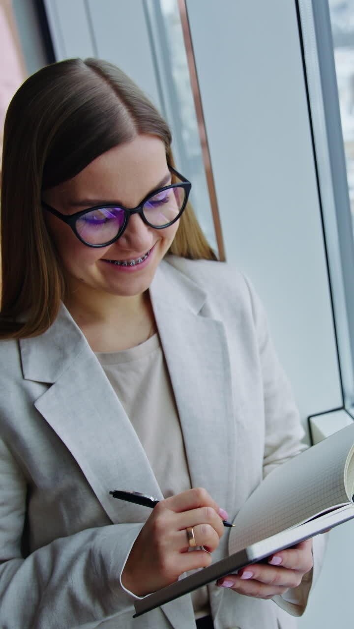 Thoughtful young woman taking notes and smiling. Female standing at the window and enjoying the cityscape. Close up. Look from above. Vertical video