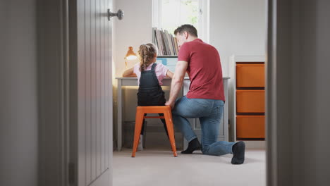 vista a través de la puerta del dormitorio mientras el padre ayuda a la hija usando la computadora portátil en el escritorio