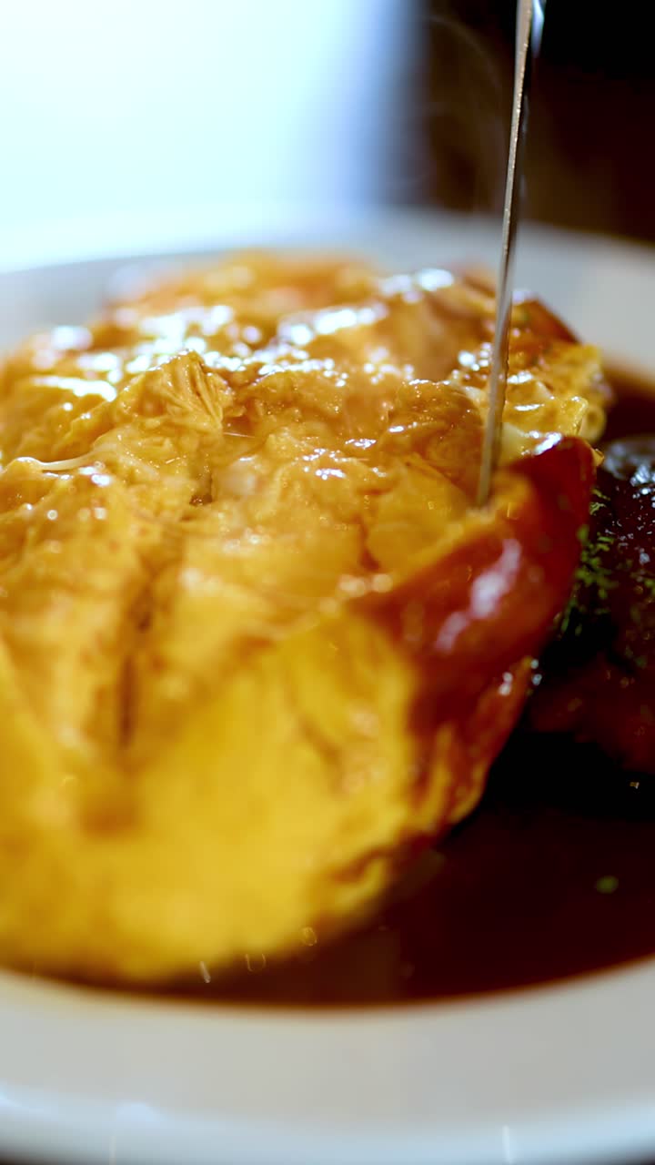Nærbilde av omurice som skjæres opp, som avslører et dampende indre ved siden av en hamburgstek i varm restaurantbelysning.