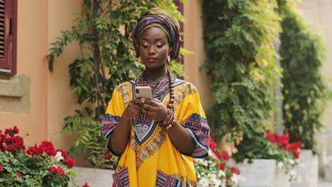 Portrait-Of-The-Young-Beautiful-Woman-In-The-Traditional-Clothes-Standing-In-The-Nice-Courtyard-With-Flowers,-Tapping-And-Texting-On-The-Smartphone,-Then-Smiling-To-The-Camera