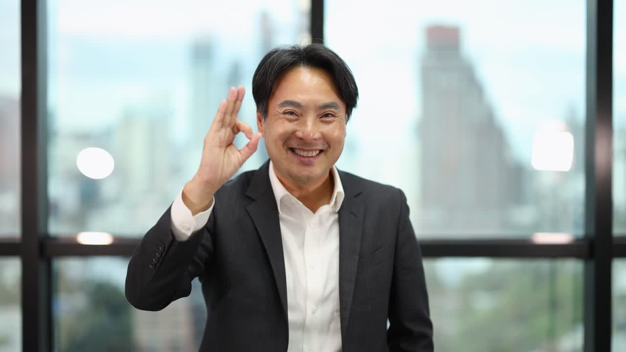 Smiling businessman in suit makes OK gesture, standing in bright modern office with city view