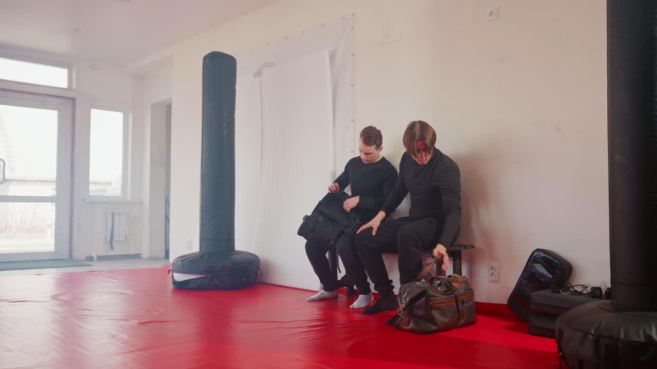 Two men inside martial arts gym with red mat, seated on wooden bench wearing black sportswear with neck tattoo, carrying brown leather bag walking by, punching bags visible in bright interior training room