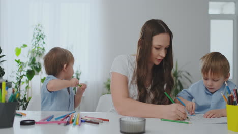 A-young-mother-with-two-children-sitting-at-a-white-table-draws-colored-pencils-on-paper-helping-to-do-homework