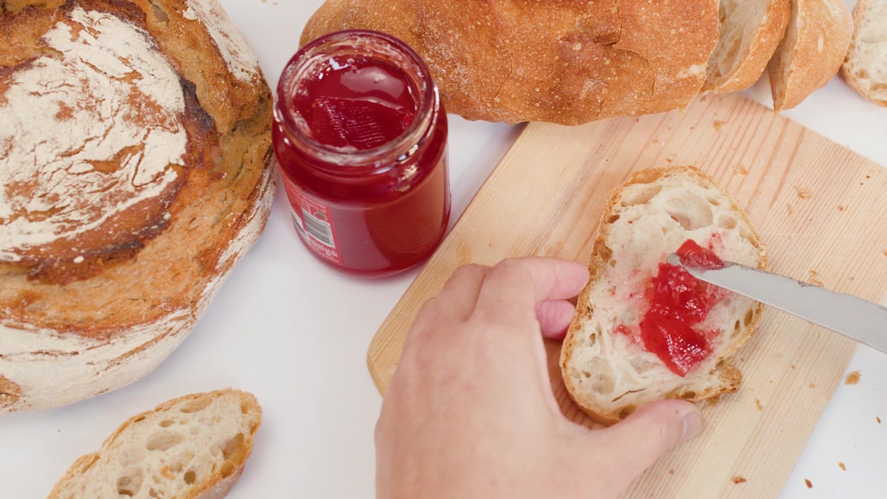 Top view of hand spreading red fruit jam on crusty bread using a knife.
