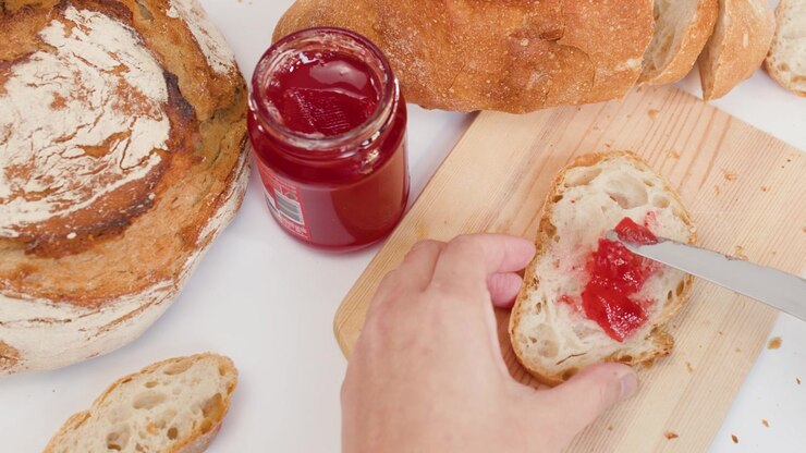 Top view of hand spreading red fruit jam on crusty bread using a knife.