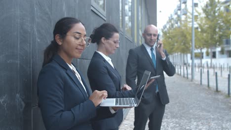 side view of business team standing at office building, using tablet, laptop and phone