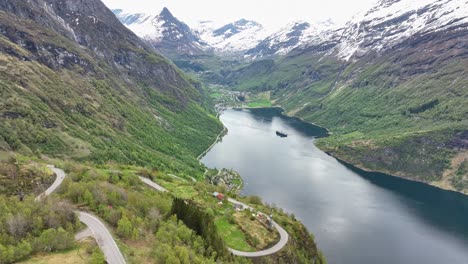 spring aerial geiranger norway - green hillsides with snow capped mountains and cruise ship on sea - ornevegen road to the left