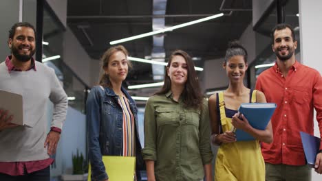 diverse group of happy work colleagues walking through corridor holding laptop, tablet and documents