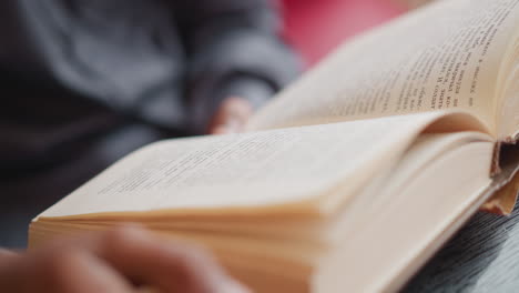 close-up of male reading book with focus on printed text and blurred hands holding pages, shirt visible in background, capturing quiet study moment, concentration, and literary engagement