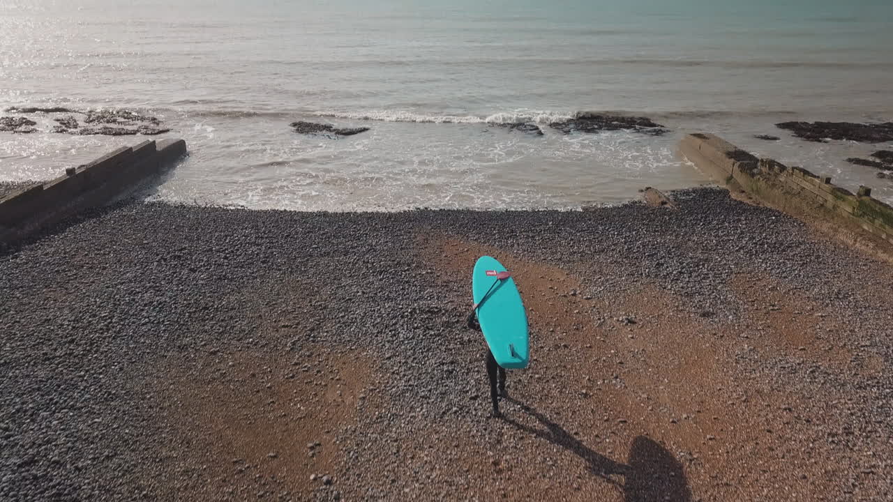 Young man walking out to sea along pebble beach, with blue paddle board balanced on his head. Drone moving forwards and camera panning up.