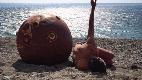 man doing yoga exercise outdoors near to an old rusty floating marine mine on the beach with rocky shore and sea background. healthy lifestyle, pollution, nature protection, war and peace concept