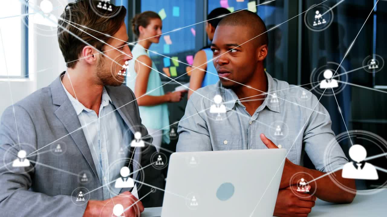 Two men sitting at table, man gesturing, laptop generating business links growing outward for team