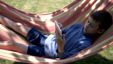 boy lies on a hammock, uses a tablet