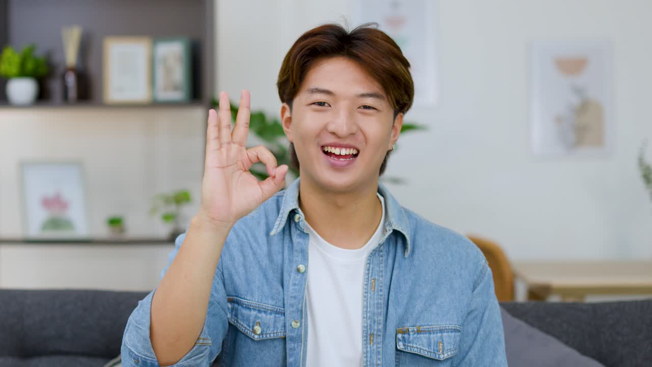 데님 셔츠를 입은 쾌활한 아시아 남성이 거실에서 좋아요 사인을 보내고 있어요.