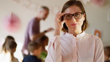 retrato de una feliz maestra que usa gafas con un peinado de bob que ajusta sus gafas y se para con confianza en una camisa rosada contra el telón de fondo de un club especial para niños para preparar a los niños para la escuela