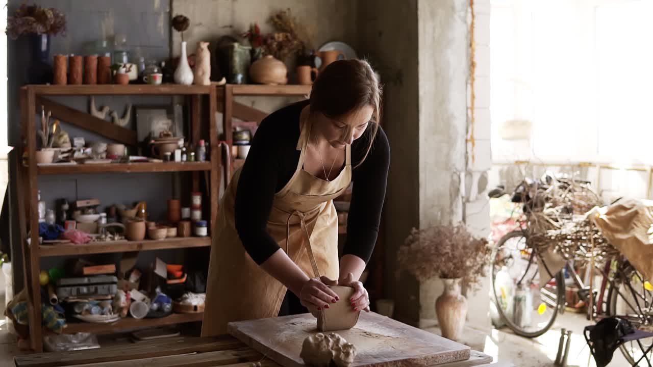 Front view of female potter wearing beige apron kneading softly clay piece on worktop, working with her hands. Pottery products on shelves behind, lens flares and sunlight from window. Slow motion