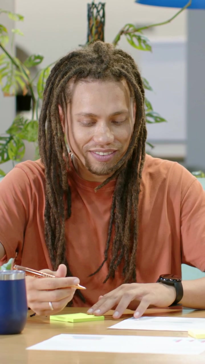 Vertical video: Scanning papers orange-shirt man pausing then writing on yellow pad during meeting