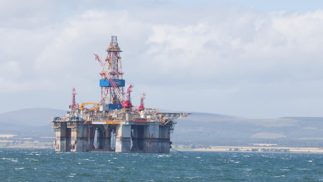 Statisk, bredt billede af offshore olieplatform i dagslys, oprørt hav og skotsk højland i det fjerne som baggrund