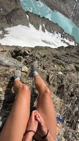 vertical 4k, young woman sitting on top of steep cliff above glacier and glacial lake, revealing shot