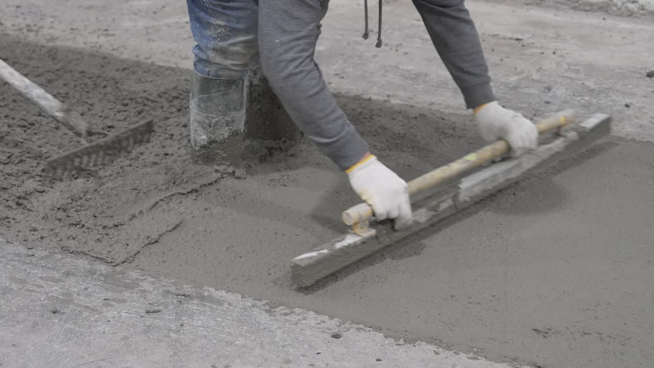 Construction worker in jeans, gloves, and boots stands knee-deep in wet concrete, leveling it with a screed and level vial. Another worker rakes cement in the background.
