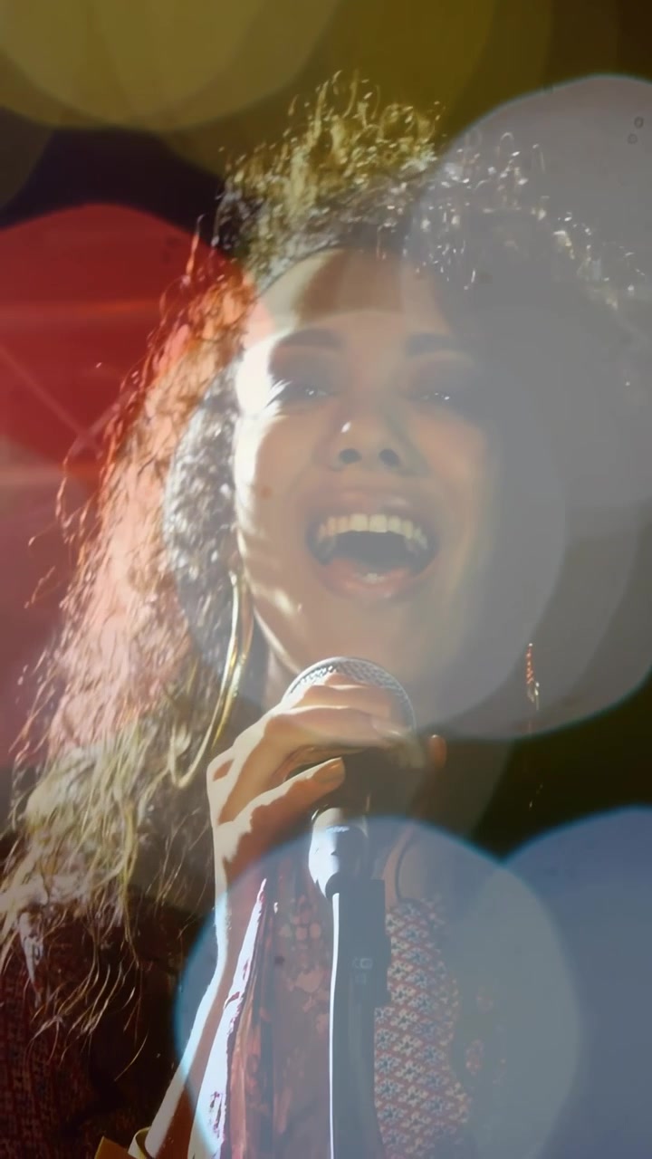 Video verticale: Cantante donna che tiene un microfono, intona una nota ed esegue musica, con bokeh fluttuante.