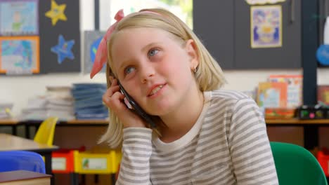 front view of caucasian schoolgirl talking on mobile phone at desk in classroom 4k