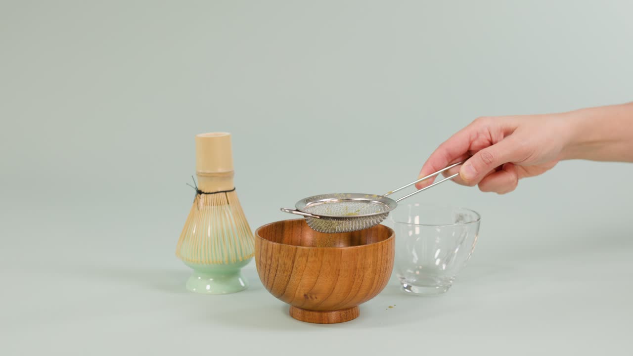 Hand sifts matcha powder through strainer into wooden bowl, with bamboo whisk and glass cup