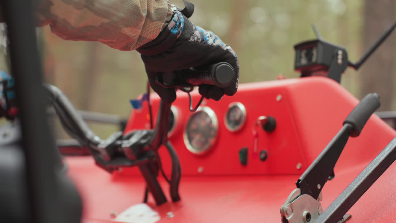 Primo piano delle mani sull'acceleratore della console di un quad rosso. Giacca mimetica e mano guantata azionano l'accensione e la leva contro lo sfondo sfocato di una pineta, creando un'atmosfera tecnica e concentrata prima di un fuoristrada estremo.