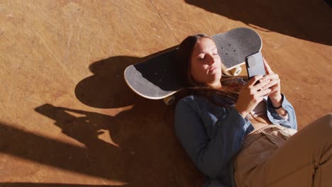 video of happy caucasian female skateboarder resting and using smartphone