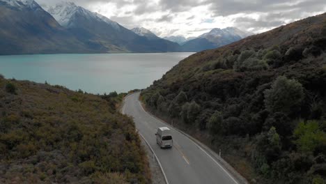 Cámara-Lenta---Autocaravana-Conduciendo-En-Las-Montañas-A-Lo-Largo-De-La-Costa-Del-Lago-Wakatipu,-Queenstown,-Nueva-Zelanda-Con-Montañas-De-Nieve-Fresca-En-El-Fondo---Drone-Aéreo