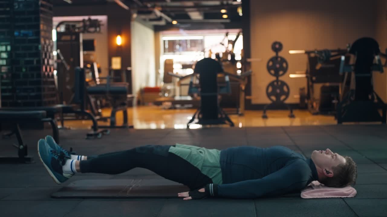 Athletic man doing leg raises lying on yoga mat, focused on abdominal muscle toning, within the vibrant dim ambiance of a modern gym - profile view
