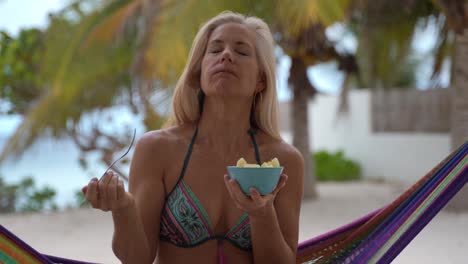 primer plano de una mujer bastante madura en bikini sentada en una hamaca en un complejo turístico, huele un pedazo de piña y luego lo gusta y sonríe a la cámara