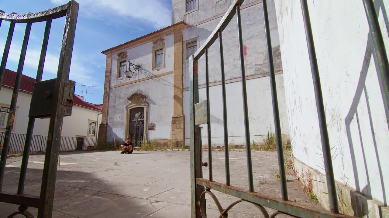 A wide shot of a man walking alone, reflecting outdoors near the historical white buildings of Coimbra, Portugal, framed by a metal gate, under a bright sunny day