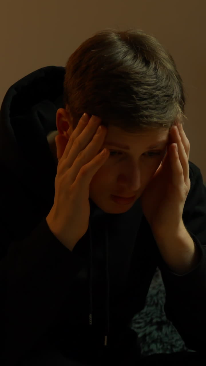 Young man burnout sitting indoors with his hands on his head, conveying stress, overwhelm, or emotional tension in a low-light setting