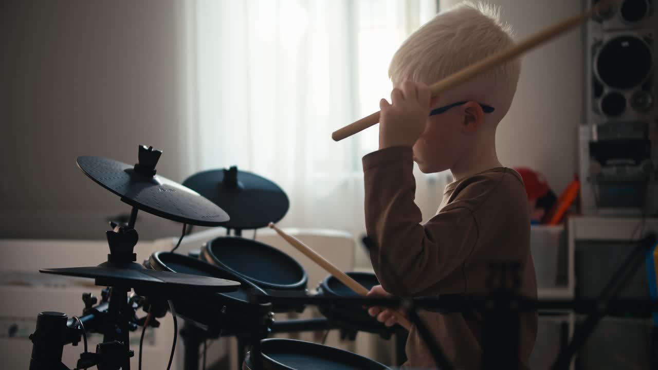 A little albino boy with white hair color in blue round glasses plays on an electronic drum set at home in his room. Happy little boy with talent for playing musical instruments holds percussion sticks in his hands and plays the drums