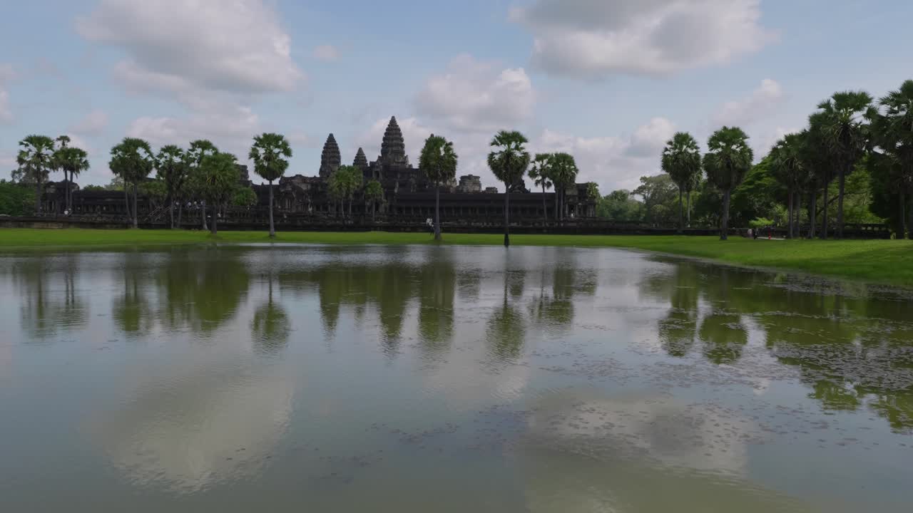 Un travelling latéral fluide en drone montrant la réflexion d’Angkor Vat dans l’étang entouré de palmiers sous un ciel lumineux avec des nuages légers