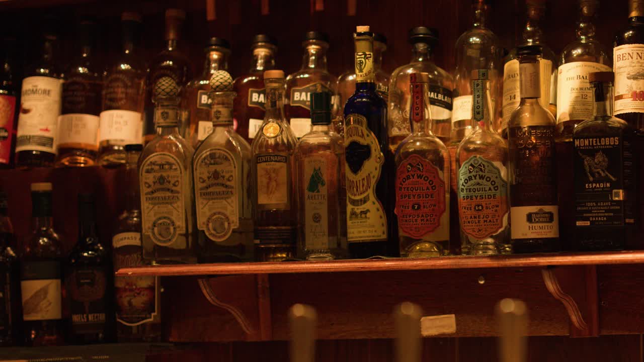 Static medium shot of various scotch and liquor bottles on wooden shelves in dim lighting