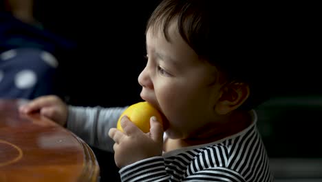 Entzückendes-8-Monate-Altes-Bengalisches-Baby,-Das-In-Eine-Orange-Beißt-Und-Mit-Einer-Hand-Auf-Dem-Tisch-Steht