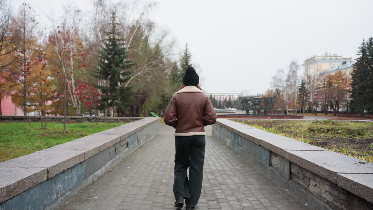 Back shot of student in black cap brown jacket walking slowly with hands in pockets on wide stone pathway through urban park surrounded by pine trees modern buildings and cloudy sky