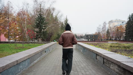 back shot of student in black cap brown jacket walking slowly with hands in pockets on wide stone pathway through urban park surrounded by pine trees modern buildings and cloudy sky