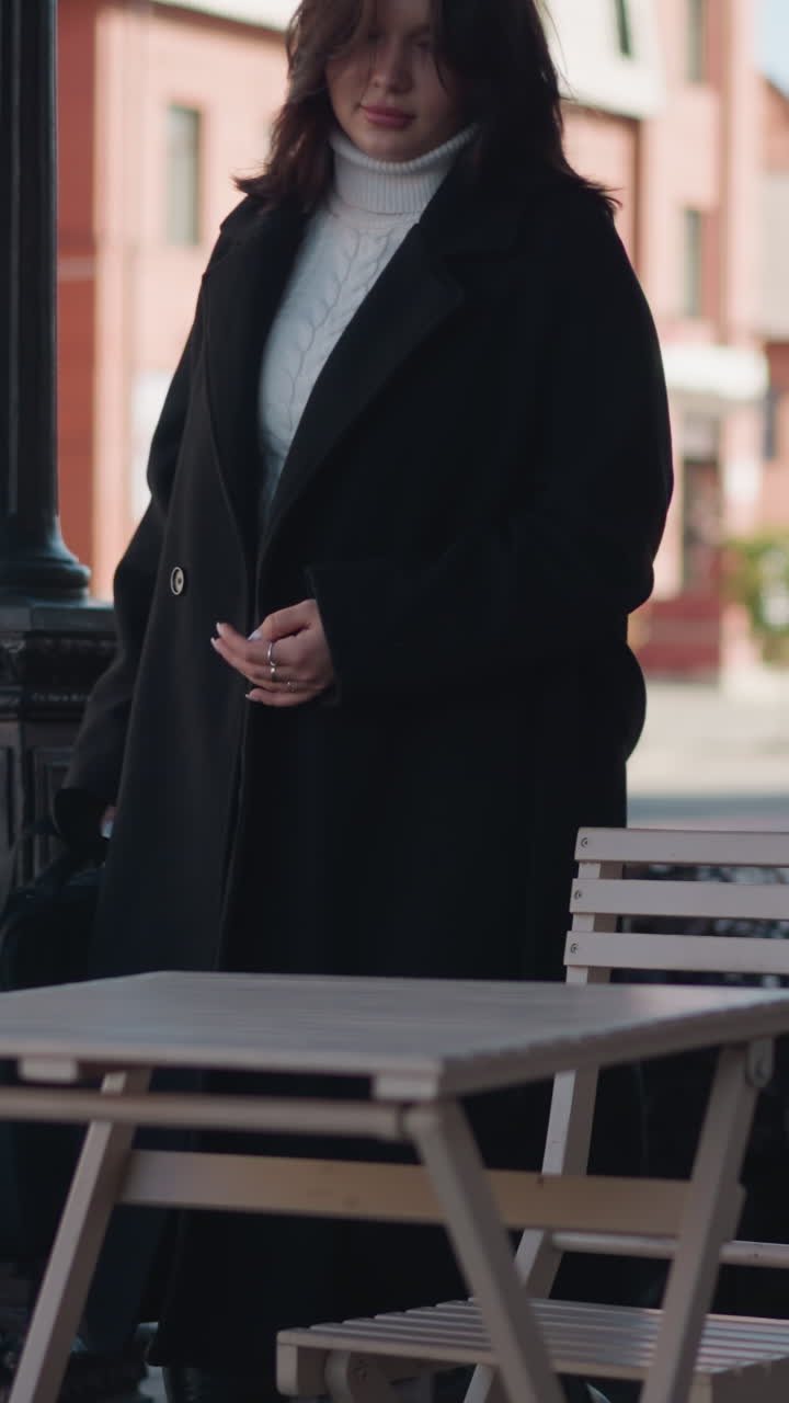 Businesswoman in black coat and white turtleneck sits down on outdoor restaurant chair, placing her bag on table with visible relief, urban background includes decorative poles, chairs and tables
