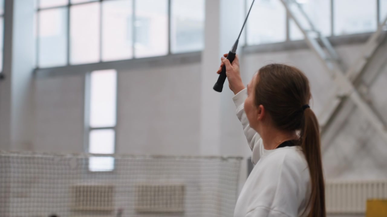 Caucasian woman practicing badminton serve indoors in spacious gymnasium, ponytail in white sweatshirt, looking up at shuttlecock, solo drill beside net, soft natural light, focused repetition