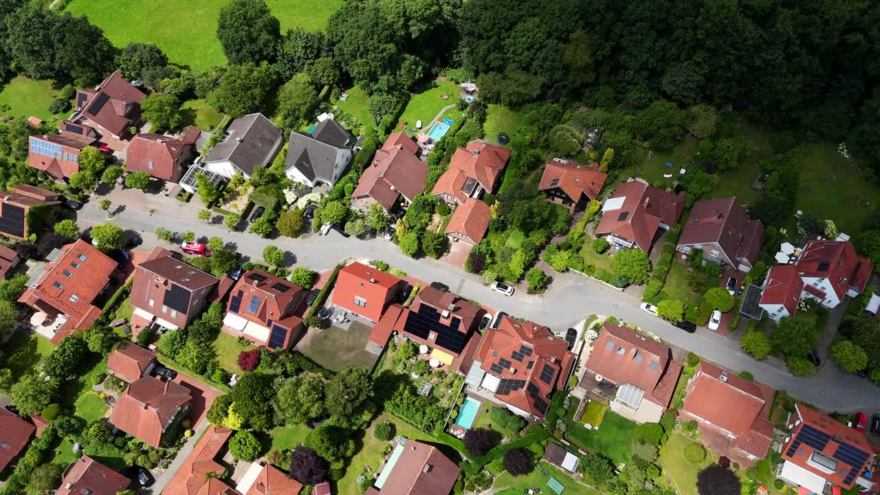 Solar panels on houses with orange tiles in suburb of town. Sunny day in summer near forest trees and path. Highway in background. Aerial top down flyover. Green renewable energy theme.