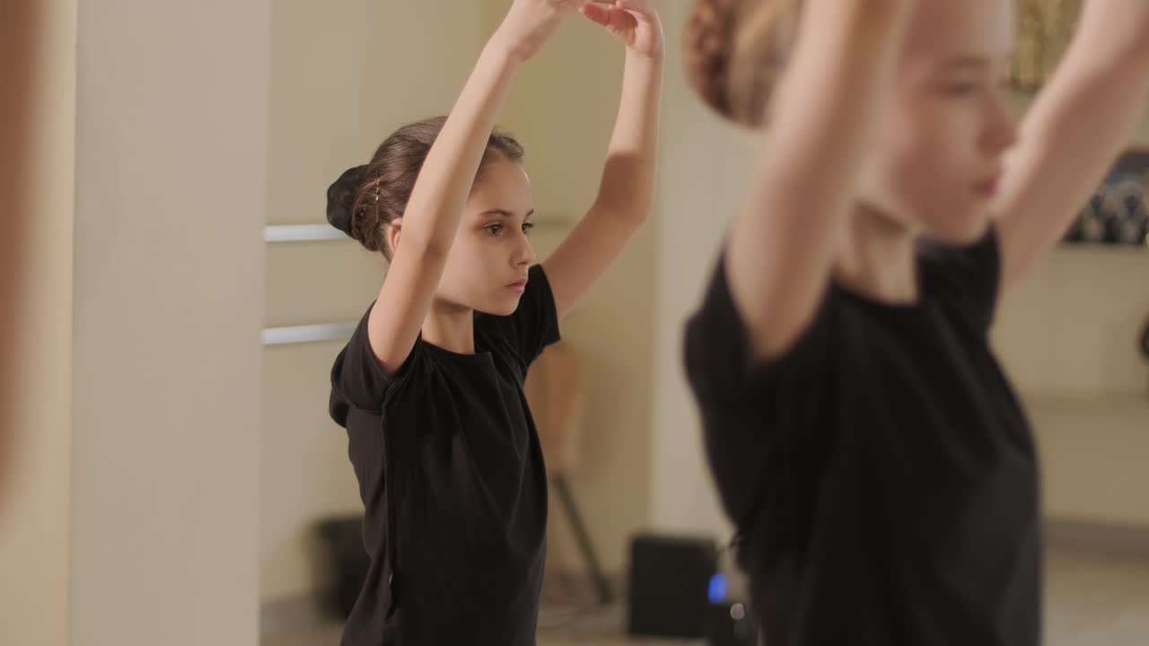 A group of young ballet students in black dancewear practicing positions in a spacious ballet studio with wooden flooring and wall-mounted barres. Focused expressions and synchronized movements.