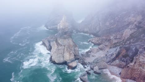 Toma-Aérea-En-órbita-De-Una-Roca-Imponente-En-La-Niebla-En-Praia-Da-Ursa,-Portugal