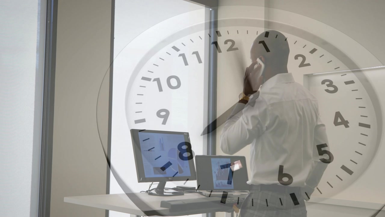 Man receiving call, standing in office holding phone, glancing at monitors, clock overlay moving