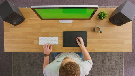 Overhead-View-Of-Male-Graphic-Designer-Or-Retoucher-Working-At-Computer-With-Green-Screen-In-Office