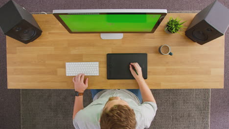 overhead view of male graphic designer or retoucher working at computer with green screen in office